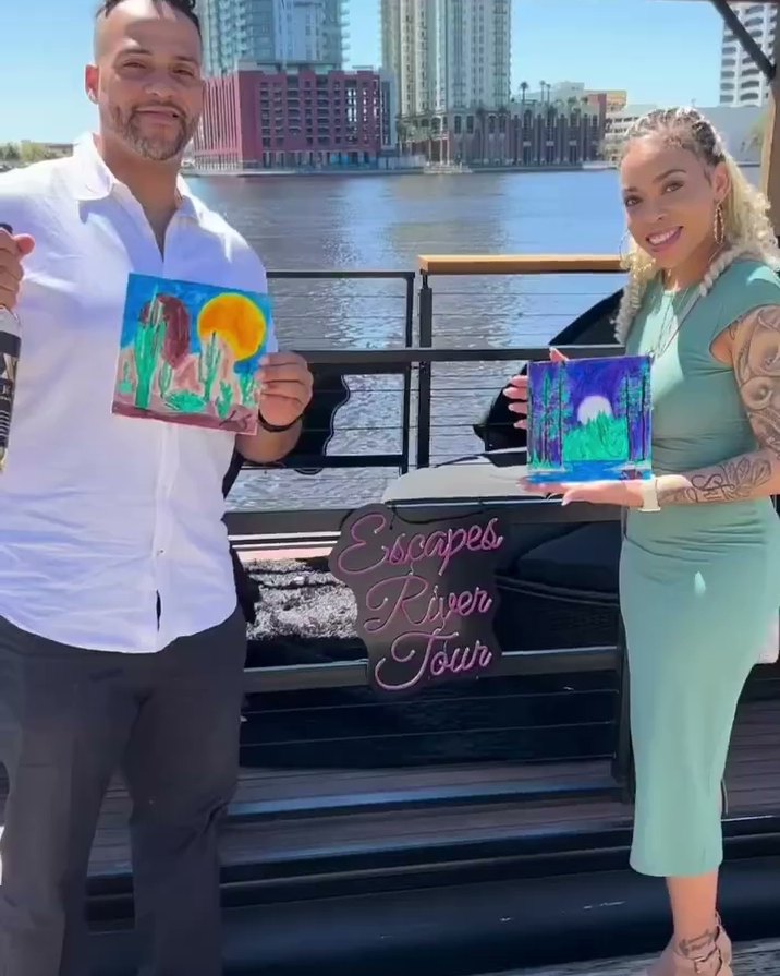 A guest holding up a finished sip-and-paint canvas beside the Escapes River Tour neon sign on the boat